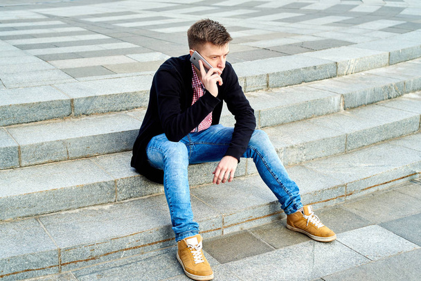 Full length body portrait of handsome male student sitting alone on stairs and talking on mobile phone, having rest in university campus, copy space. Education, rest and relax, travel and tourism concept - Фото, зображення