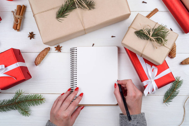 Γυναίκα γράψιμο τους στόχους του νέου έτους (ή να κάνει τον κατάλογο) στο κενό σημειωματάριο στο γραφείο με διακοσμήσεις Χριστουγέννων, κουτιά δώρων, κλαδιά δέντρων ελάτης και κώνους, αντίγραφο χώρου. Το Top view, επίπεδη lay  - Φωτογραφία, εικόνα
