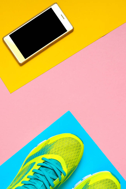 Пара спортивной обуви и мобильный телефон на красочном фоне. Новые кроссовки на розовом, синем и желтом фоне, скопировать пространство. Накладные расходы на кроссовки и смартфон. Вид сверху, плоский
 - Фото, изображение