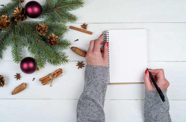 Top view of Christmas background with blank empty diary and girl's hands with pen writing to do list on white wooden background, free space. Flat lay, top view - Zdjęcie, obraz