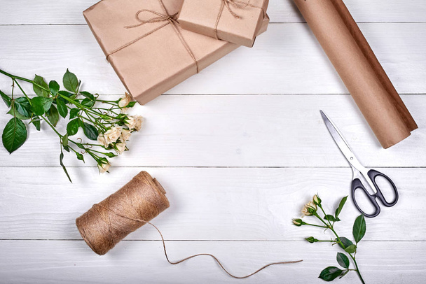 Gift wrapping background. Rolls of kraft wrapping paper, rope, branch of roses, gift boxes and scissors on wooden background, free space. Flat lay, top view  - Photo, image
