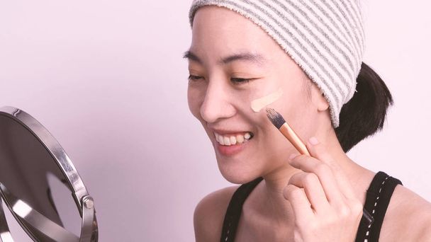 Asyalı kız ya da kadın 40 yaşında Japon görünümlü güzel bir kız cildinin kusursuz ve gerçek görünmesine yardım ettiği için hassas cildindeki sıvı ve kozmetik fırçayla makyaj yapıyor. - Fotoğraf, Görsel