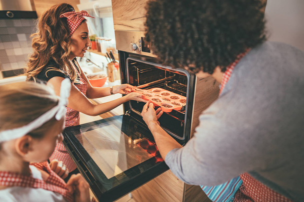Happy parents and daughter putting cookies in oven to bake - Photo, Image