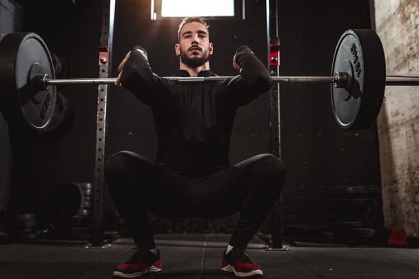 Young muscular man doing squat exercise with barbell at gym - Photo, image