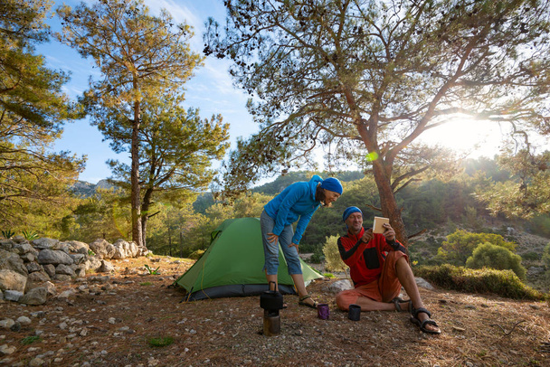 Couple of happy travelers is drinking coffee in the mountains among lush pines at sunny morning and using tablet pc - camp during adventure travel along Lycian way, Turkey. Backlight, wide angle. - Zdjęcie, obraz