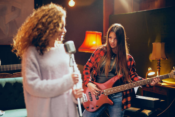 zwei Frauen üben für den Auftritt. Frau mit lockigem Haar hält Mikrofon in der Hand und singt, während eine andere Bassgitarre spielt. Heimstudio. - Foto, Bild