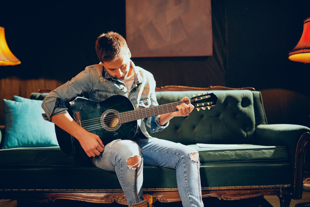 Young bearded focused Caucasian man sitting on the sofa and playing acoustic guitar. Home studio interior. - Photo, Image