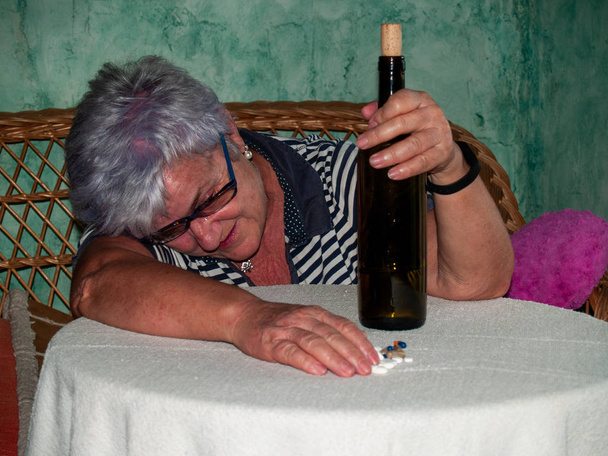 Una mujer mayor con una botella de vino y un gran número de pastillas en la mano de diferentes colores y tamaños, acostada en una mesa
 - Foto, Imagen