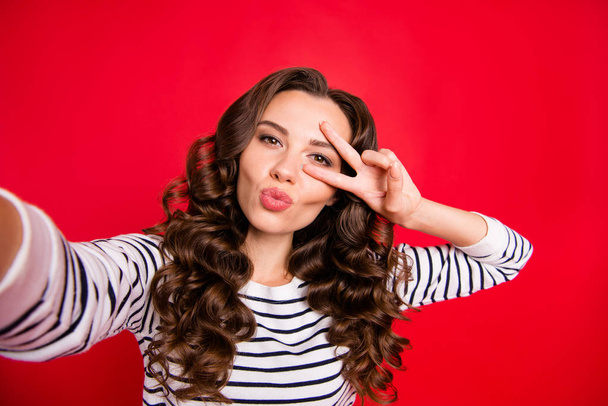 Self-portrait of nice cute lovely sweet pretty attractive positive cheerful wavy-haired lady wearing striped pullover showing v-sign near eye isolated over bright vivid shine red background - Фото, зображення