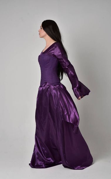 portrait pleine longueur de belle fille aux longs cheveux noirs, portant une robe médiévale fantaisie violette. pose debout sur fond de studio gris
. - Photo, image