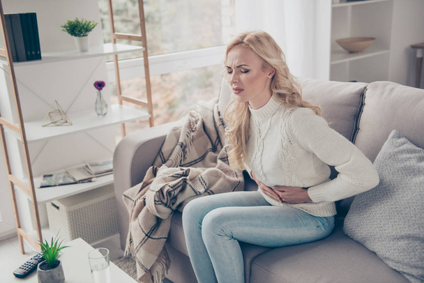 Attractive stylish gloomy wavy-haired lady wife housewife in sweater sitting on divan writhing in pain early term pregnancy digestion problem trouble in light white interior room - 写真・画像
