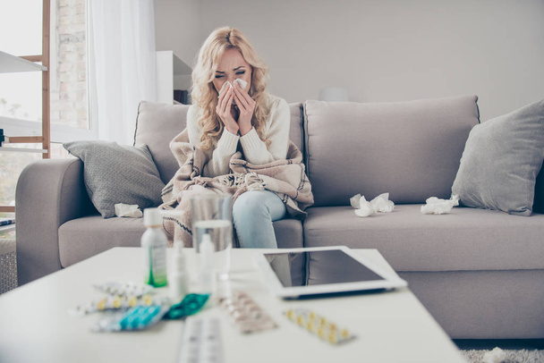 Attractive wavy-haired girl sitting on divan wrapped in blanket in front of pile medicine colorful pills c-vitamin health support suffering healing allergy in light room - Foto, imagen