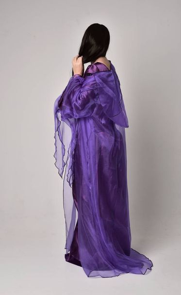 retrato de comprimento total da menina bonita com cabelo preto longo, vestindo vestido medieval fantasia roxa e capa. em pé pose no fundo do estúdio cinza
. - Foto, Imagem