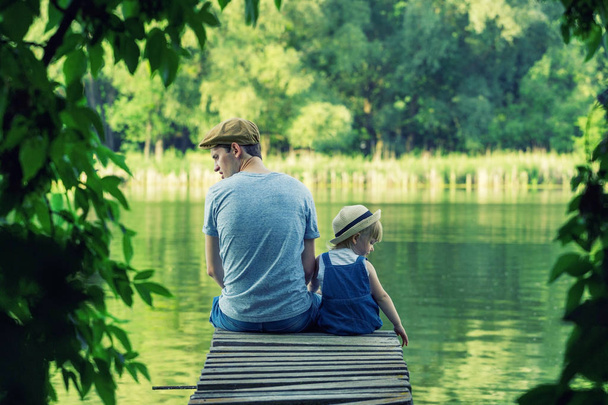 Отец и маленькая девочка в комбинезоне сидят на мосту и смотрят на разные стороны. Вид сзади
. - Фото, изображение