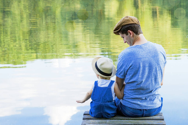 Dad and a tiny girl in dungarees relax at the river. Back view, copy space. - Zdjęcie, obraz