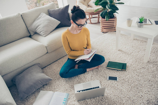 Above high angle view of nice lovely attractive minded serious focused concentrated girl with hair bun sitting in lotus position barefoot on carpet writing courses materials in light interior room - Photo, image