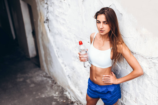 Pretty sporty girl standing near wall after training with bottle of water - Photo, image