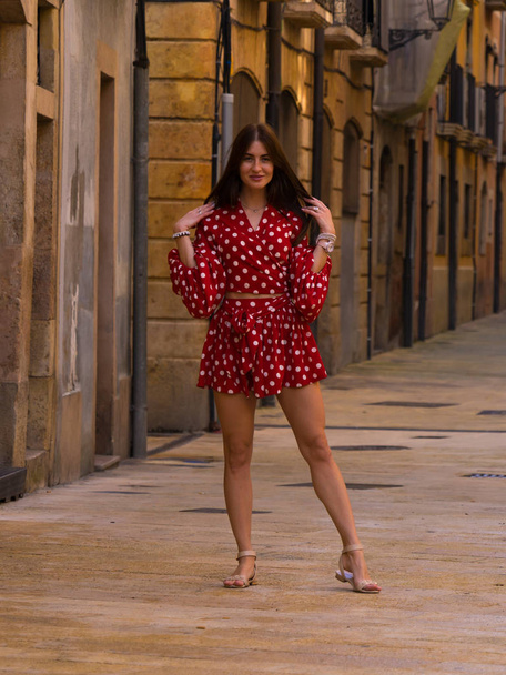 Spain vacation series. Narrow street in Tarragona. Young girl in red dress in white peas near the yellow brick wall and paved sidewalk on subject of travel, summer, vacation,  youth, beauty. - Photo, image