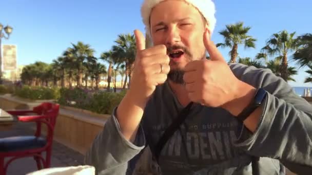 Porträt eines positiven Mannes mit Weihnachtsmütze im Strandcafé mit Palmen und Meer - Filmmaterial, Video