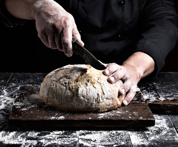 muž krájí nožem kulatý bochník bílé pšeničné mouky na hnědé dřevěné desce - Fotografie, Obrázek