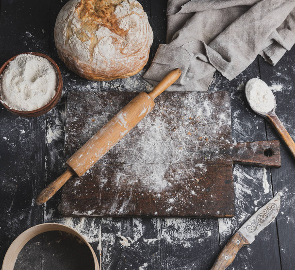 baked bread, white wheat flour, wooden rolling pin and old cutting board on a black table, top view - Fotoğraf, Görsel