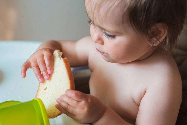 Dziewczynka siedzi w krzesełko i jedzenie kawałek chleba. Słodkie dziecko. Szczęśliwe dziecko - Zdjęcie, obraz