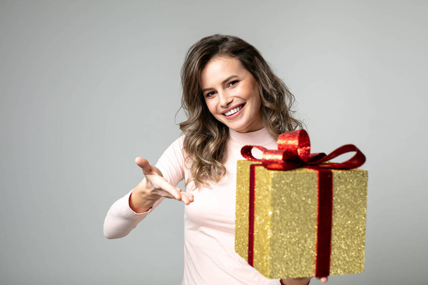 beautiful woman in pink sweater presenting her golden gift to camera on gray background - Zdjęcie, obraz