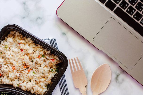 köstlicher gebratener Reis in Lunchbox auf Serviette mit Kochlöffel und Gabel und Computer-Laptop am Arbeitsplatz für Fertiggerichte und Lebensmittelkonzept - Foto, Bild