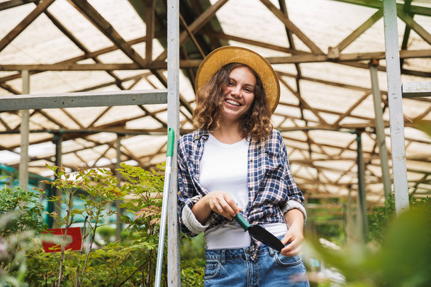 Portrait of young happy cheerful florist woman 20s posing and working in greenhouse over plants - Φωτογραφία, εικόνα