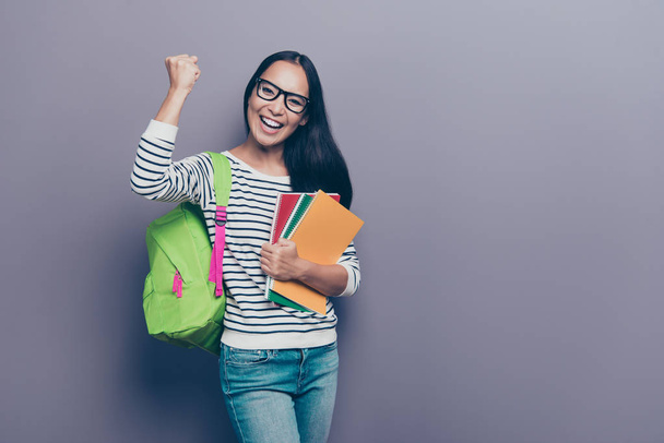 Portrait of attractive cheerful straight-haired brunette girl in striped pullover eyeglasses eyewear holding in hand colorful book school green bag waving isolated over gray pastel violet background - Fotó, kép