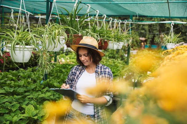 Pretty young woman working in a greenhouse, writing notes in a tablet - 写真・画像