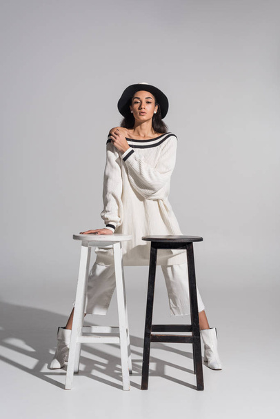 attractive african american girl in stylish white clothes and hat standing near black and white chairs on white - 写真・画像