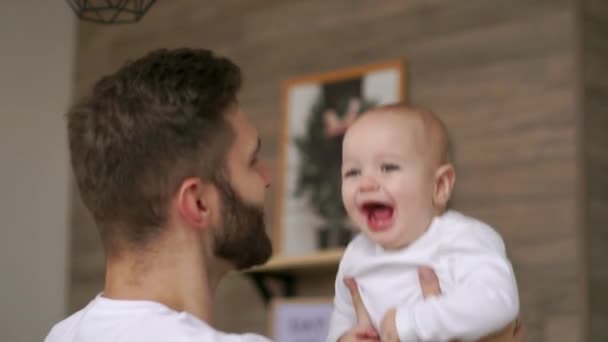 Father plays with infant son in white t-shirt looks at him and laughs. Laughing baby looking at the camera. Loving father - Záběry, video
