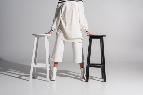 cropped image of girl in fashionable sweater and trousers standing between black and white chairs on white - Foto, Imagem