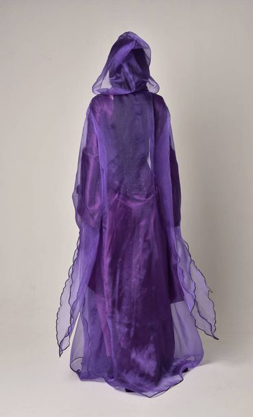 retrato de longitud completa de hermosa chica con el pelo largo y negro, usando vestido medieval de fantasía púrpura y capa. pose de pie sobre fondo gris estudio
. - Foto, Imagen