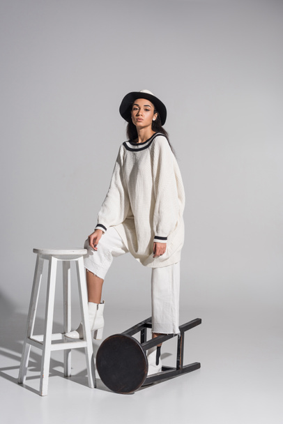attractive african american girl in stylish white clothes and hat standing on black and white chairs on white - Photo, Image