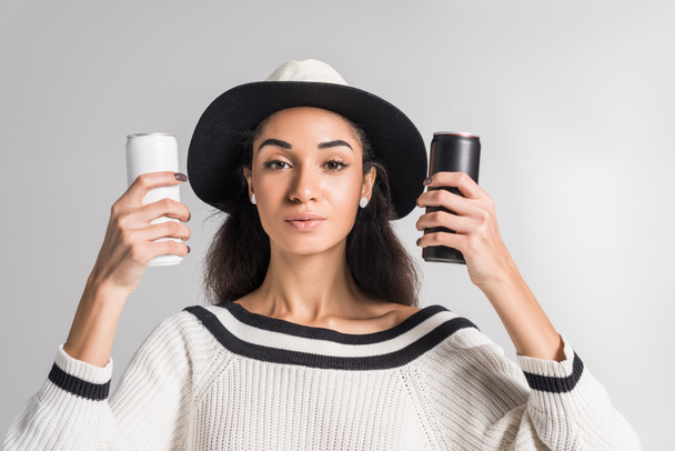 attractive african american girl in stylish white sweater and hat holding white and black cans and looking at camera isolated on white - Фото, изображение