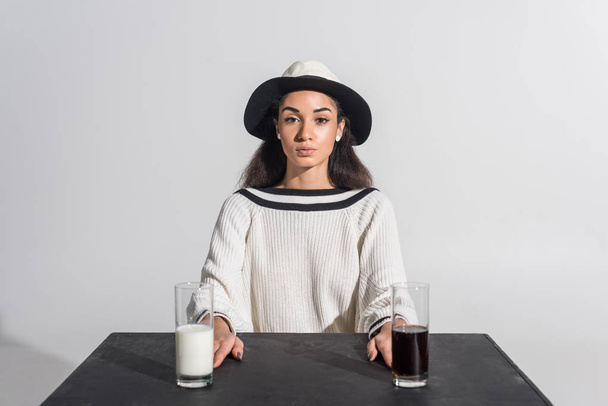 beautiful african american woman in stylish white clothes and hat sitting at black table with milk and soda in glasses on white - Foto, immagini