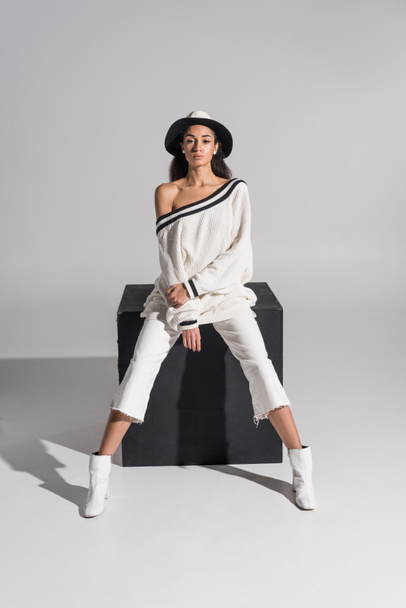 attractive african american girl in stylish white clothes and hat sitting on black cube and looking at camera on white - Foto, Imagen