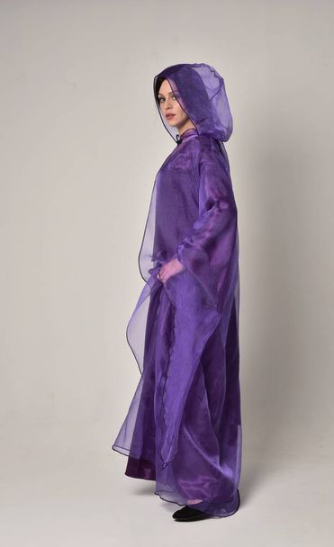 retrato de longitud completa de hermosa chica con el pelo largo y negro, usando vestido medieval de fantasía púrpura y capa. pose de pie sobre fondo gris estudio
. - Foto, imagen