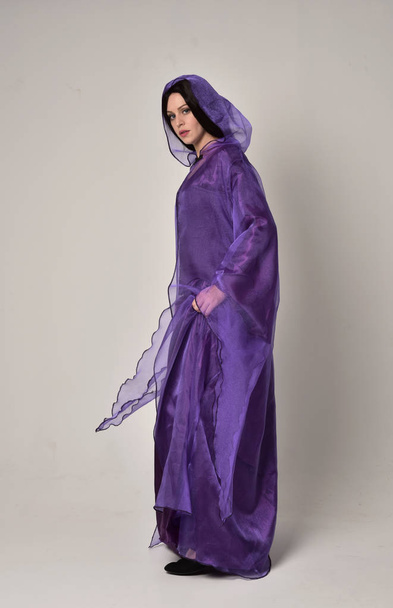 full length portrait of beautiful girl with long black hair,   wearing purple fantasy medieval gown and cloak. standing pose on grey studio background. - Фото, изображение