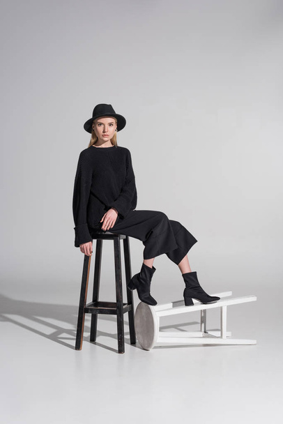 attractive caucasian blonde woman in black clothes and hat sitting on chair and looking at camera on white - 写真・画像