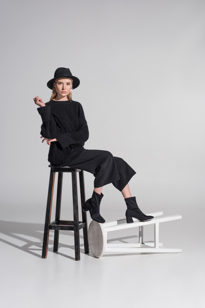 beau modèle blond caucasien dans des vêtements noirs élégants et chapeau assis sur chaise sur blanc
 - Photo, image