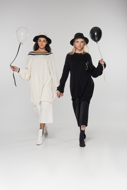 όμορφη πολυπολιτισμική ζευγάρι λεσβιών με μαύρα και άσπρα ρούχα, κρατώντας τα χέρια και το περπάτημα με δύο μπαλόνια σε λευκό - Φωτογραφία, εικόνα