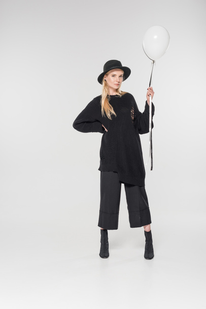 attractive caucasian blonde woman in stylish black clothes and hat holding balloons on white and looking at camera - Fotó, kép