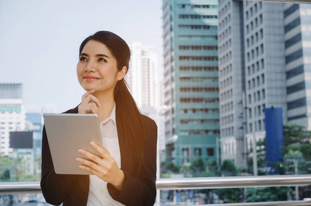 beautiful smiling young asian investor business woman wearing modern black suit thinking and looking up with mobile tablet in modern city, investment,  network technology, internet, digital concept  - Zdjęcie, obraz