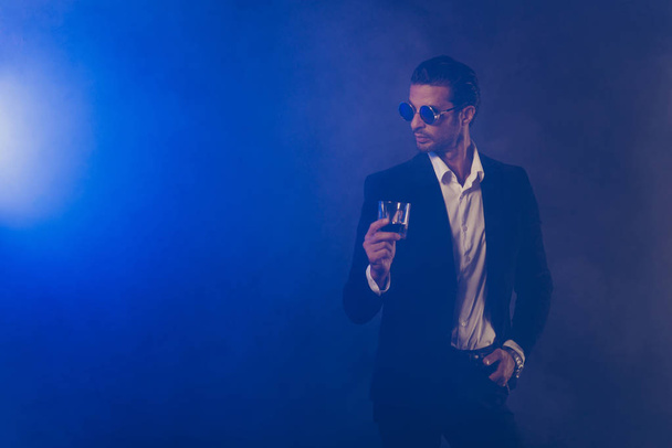 Portrait of nice fashionable attractive handsome man wearing black jacket round eyewear eyeglasses holding wine glass isolated over dark blue fume smoke background - Foto, Imagem