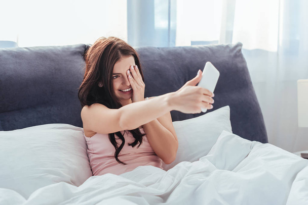 smiling girl taking selfie on smartphone while sitting on bed during morning time at home - Fotoğraf, Görsel