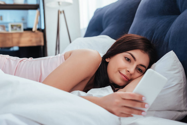 cheerful pretty girl using smartphone on bed during morning time at home - Fotoğraf, Görsel