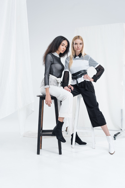 attractive multiethnic women in black and white clothes posing on chairs and looking at camera on white - Valokuva, kuva